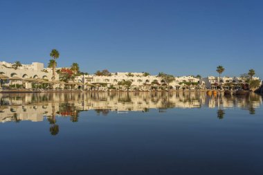 Gündoğumunda, Mısır 'ın Sharm El Sheikh tatil beldesindeki sakin deniz suyuna binalar, güneşlikler ve şemsiyeler yansıtılır.
