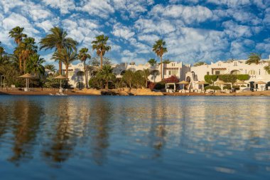 Gündoğumunda, Mısır 'ın Sharm El Sheikh tatil beldesindeki sakin deniz suyuna binalar, güneşlikler ve şemsiyeler yansıtılır.