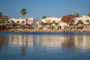 Gündoğumunda, Mısır 'ın Sharm El Sheikh tatil beldesindeki sakin deniz suyuna binalar, güneşlikler ve şemsiyeler yansıtılır.