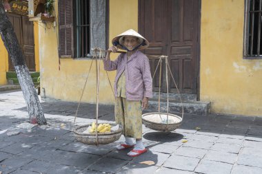 Hoi An, Vietnam - 1 Temmuz 2020: Vietnam 'da Hoi An, Vietnam' daki eski sarı duvarın arkasında elinde meyve sepeti olan hasır şapkalı Vietnamlı kadın