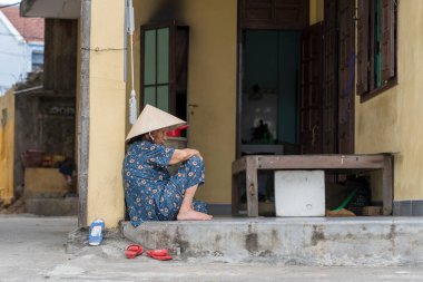 Hoi An, Vietnam - 16 Mart 2020: Hasır şapkalı yaşlı Vietnamlı kadın Hoi An, Vietnam 'daki evinin yakınındaki bir sokakta oturuyor
