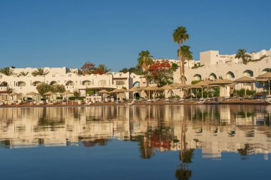 Gündoğumunda, Mısır 'ın Sharm El Sheikh tatil beldesindeki sakin deniz suyuna binalar, güneşlikler ve şemsiyeler yansıtılır.