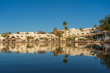 Gündoğumunda, Mısır 'ın Sharm El Sheikh tatil beldesindeki sakin deniz suyuna binalar, güneşlikler ve şemsiyeler yansıtılır.