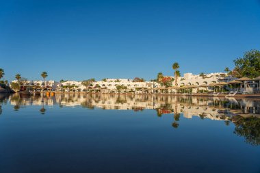 Gündoğumunda, Mısır 'ın Sharm El Sheikh tatil beldesindeki sakin deniz suyuna binalar, güneşlikler ve şemsiyeler yansıtılır.