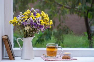 Eski bir kitap, renkli bir buket çiçek ve arka planda beyaz bir fincan çay. Yaz günü bahçede yağmur damlaları var. Kapatın, boşluğu kopyalayın.
