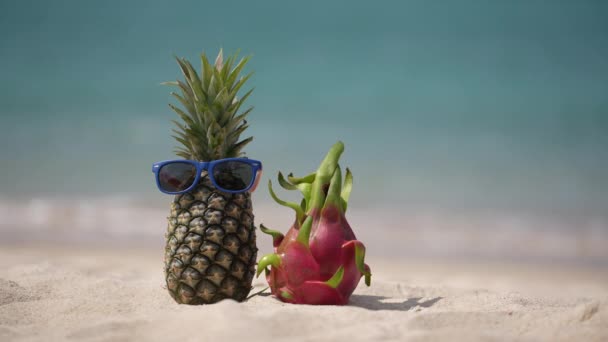 ターコイズブルーの海の水に対して砂の熱帯のビーチでサングラスとピンクのドラゴンフルーツで魅力的なパイナップルを熟す 夏休みのコンセプト — ストック動画