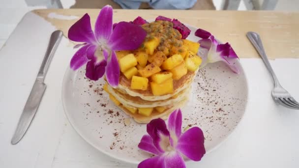 カフェのテーブルの上のプレートにトロピカルフルーツとおいしいパンケーキ クローズアップ 健康的な朝食 マンゴー ドラゴンフルーツ パッションフルーツの美味しいパンケーキ 蘭の花で飾られています — ストック動画