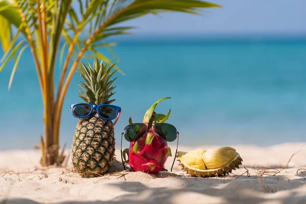 Ripe attractive pineapple and pink dragon fruit in sunglasses and fresh durian on the sand tropical beach against turquoise sea water, Thailand. Summer vacation concept