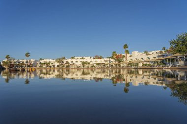 Gündoğumunda, Mısır 'ın Sharm El Sheikh tatil beldesindeki sakin deniz suyuna binalar, güneşlikler ve şemsiyeler yansıtılır.