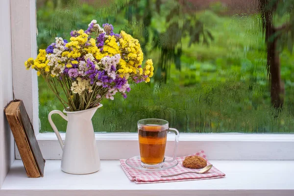 Eski bir kitap, renkli bir buket çiçek ve arka planda beyaz bir fincan çay. Yaz günü bahçede yağmur damlaları var. Kapatın, boşluğu kopyalayın.