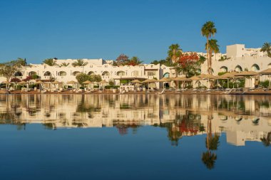 Gündoğumunda, Mısır 'ın Sharm El Sheikh tatil beldesindeki sakin deniz suyuna binalar, güneşlikler ve şemsiyeler yansıtılır.