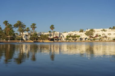 Gündoğumunda, Mısır 'ın Sharm El Sheikh tatil beldesindeki sakin deniz suyuna binalar, güneşlikler ve şemsiyeler yansıtılır.