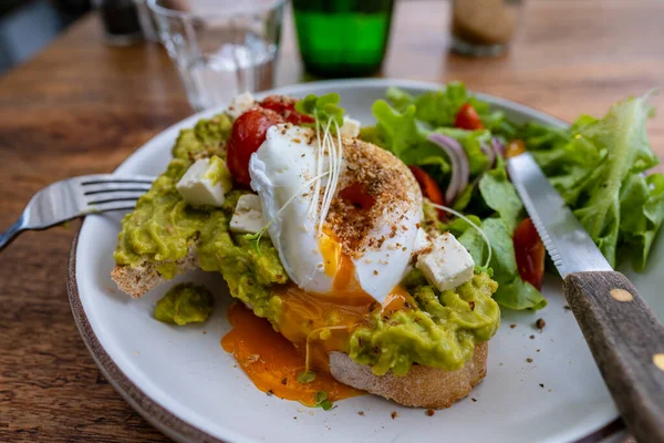Sourdough toast, poached eggs, avocado pulp and fresh vegetables on plate in cafe, close up, breakfast time