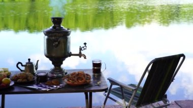 Ukrayna 'nın yeşil ormanlarında sakin su gölü yakınındaki masada beyaz duman ve yiyecek bulunan klasik metal çay semaverleri. Doğa gıda konsepti
