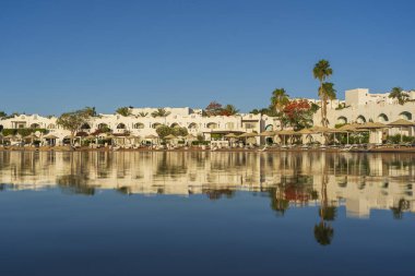 Gündoğumunda, Mısır 'ın Sharm El Sheikh tatil beldesindeki sakin deniz suyuna binalar, güneşlikler ve şemsiyeler yansıtılır.