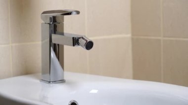 Modern banyodaki bej kaplamalı musluktan akan su ile birlikte beyaz seramik bidet detayları. Basit stil ve modern ev dekorasyonu. Kapat.