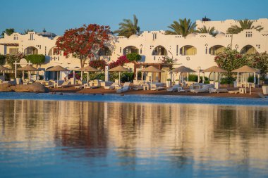 Gündoğumunda, Mısır 'ın Sharm El Sheikh tatil beldesindeki sakin deniz suyuna binalar, güneşlikler ve şemsiyeler yansıtılır.