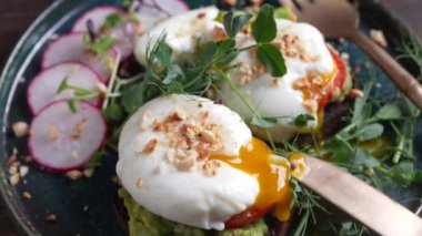 Bread toast, poached eggs, avocado pulp, pea microgreens  and fresh vegetables on plate in cafe, close up, rotates, breakfast time