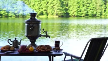 Ukrayna 'nın yeşil ormanlarında sakin su gölü yakınındaki masada beyaz duman ve yiyecek bulunan klasik metal çay semaverleri. Doğa gıda konsepti