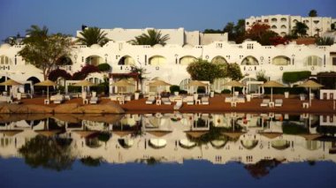 Gündoğumunda, Mısır 'ın Sharm El Sheikh tatil beldesindeki sakin deniz suyuna binalar, güneşlikler ve şemsiyeler yansıtılır.
