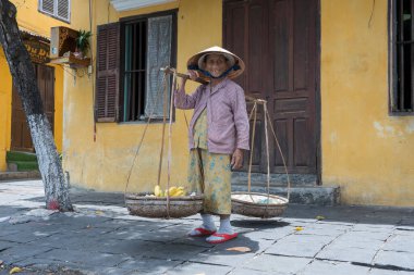 Hoi An, Vietnam - 1 Temmuz 2020: Vietnam 'da Hoi An, Vietnam' daki eski sarı duvarın arkasında elinde meyve sepeti olan hasır şapkalı Vietnamlı kadın