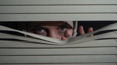 Kapalı alandaki başörtülü esmer kadının ön portresini kapat geceleri elleriyle pencereye panjur iterek. Dışarıdan korkunç görünüyor.