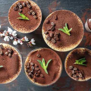 Geleneksel İtalyan tatlı tiramisusu kakao tozu, kahve çekirdeği ve yeşil nane şekeri ile süslenmiş, yakın plan, üst manzara. Çeşitli kadehlerdeki tiramisu çeşitleri siyah arka planda.