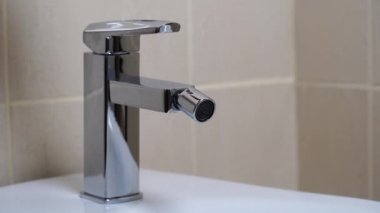 Modern banyodaki bej kaplamalı musluktan akan su ile birlikte beyaz seramik bidet detayları. Basit stil ve modern ev dekorasyonu. Kapat.