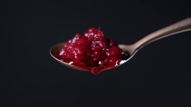 Kırmızı ahududu reçelli kaşık ve bir damla. Kırmızı ahududu reçeli siyah arka planda metal kaşıktan damlıyor ve aşağı akıyor. Yaklaş. Ev yapımı kırmızı ahududu reçelli pirinç kaşık.