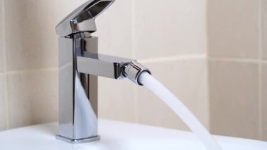Modern banyodaki bej kaplamalı musluktan akan su ile birlikte beyaz seramik bidet detayları. Basit stil ve modern ev dekorasyonu. Kapat.
