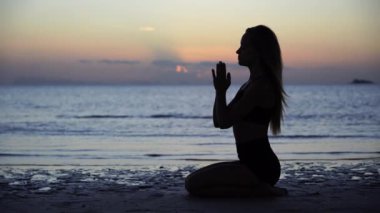 Gün batımında tropik plajda yoga pozunda oturan kadın silueti. Beyaz kız deniz suyunun yanında yoga yapıyor..