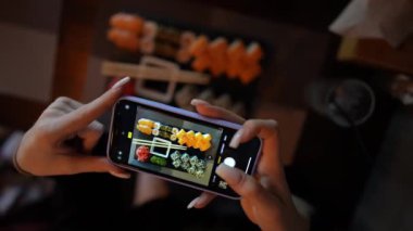 Kız, rahat Japon restoranında akıllı telefon kullanarak suşi fotoğrafı çekiyor. Kadın eli, modern akıllı telefonla yiyeceklerin fotoğrafını çeker. Yaklaş, 4K