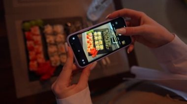Kız, rahat Japon restoranında akıllı telefon kullanarak suşi fotoğrafı çekiyor. Kadın eli, modern akıllı telefonla yiyeceklerin fotoğrafını çeker. Yaklaş, 4K