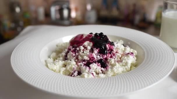 Fresh homemade cottage cheese with sour cream and blueberry jam in a white plate rotates on the table, close up. Delicious morning breakfast