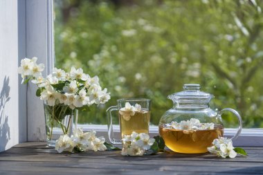 Yaz günü bahçenin yanındaki pencere eşiğinde nefis sıcak bitki çayı ve güzel bir buket yasemin çiçeği, yakın çekim. Cam çaydanlık ve fincanda sıcak yasemin çayı