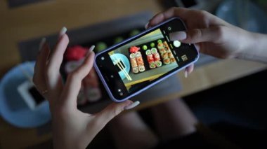 Kız, rahat Japon restoranında akıllı telefon kullanarak suşi fotoğrafı çekiyor. Kadın eli, modern akıllı telefonla yiyeceklerin fotoğrafını çeker. Yaklaş, 4K