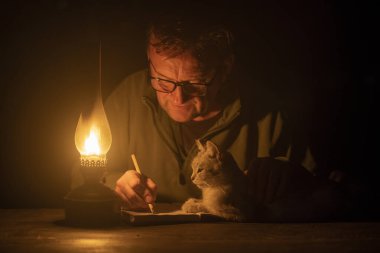 Adam defterine kurşun kalemle gaz lambasının ışığıyla yazıyor ve yakınlarda bir kedi yatıyor, gece vakti, kapat.