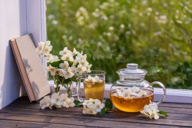 Yaz günü bahçenin yanındaki pencere eşiğinde nefis sıcak bitki çayı ve güzel bir buket yasemin çiçeği, yakın çekim. Cam çaydanlık ve fincanda sıcak yasemin çayı