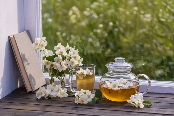 庭の近くの夏の日に自宅の窓辺でおいしいホットハーブティーとジャスミンの花の美しい花束を閉じます グラスティーポットとカップで熱々のジャスミン茶 ストック写真