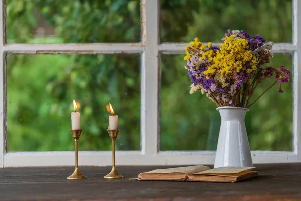 Two burning candles in a bronze candlestick an open old book and a bouquet of colorful flowers on the windowsill next to the window in the evening, close up , indoors