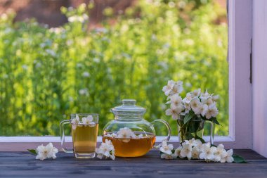 Yaz günü bahçenin yanındaki pencere eşiğinde nefis sıcak bitki çayı ve güzel bir buket yasemin çiçeği, yakın çekim. Cam çaydanlık ve fincanda sıcak yasemin çayı