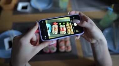 Kız, rahat Japon restoranında akıllı telefon kullanarak suşi fotoğrafı çekiyor. Kadın eli, modern akıllı telefonla yiyeceklerin fotoğrafını çeker. Yaklaş, 4K