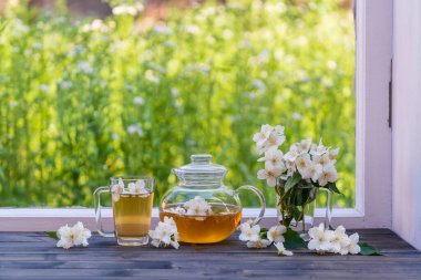Yaz günü bahçenin yanındaki pencere eşiğinde nefis sıcak bitki çayı ve güzel bir buket yasemin çiçeği, yakın çekim. Cam çaydanlık ve fincanda sıcak yasemin çayı