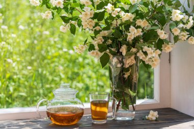 Yaz günü bahçenin yanındaki pencere eşiğinde nefis sıcak bitki çayı ve güzel bir buket yasemin çiçeği, yakın çekim. Cam çaydanlık ve fincanda sıcak yasemin çayı
