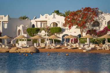 Gündoğumunda, Mısır 'ın Sharm El Sheikh tatil beldesindeki sakin deniz suyuna binalar, güneşlikler ve şemsiyeler yansıtılır.