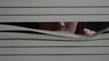 Close up front portrait from indoors of young girl pushing blinds at window with hands. Looking fearfully outside at night