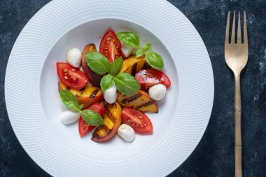 Peynirli ızgara şeftali salatası, domates, yeşil fesleğen ve sos. Siyah arka planda beyaz tabakta taze gurme salatası, yakın plan, üst görünüm