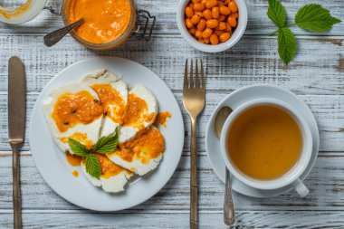 Slices of adyghe cheese poured over sea buckthorn jam with green tea and fresh raw sea buckthorn berries in white cup during breakfast on wooden table, close-up, top view. Healthy food concept