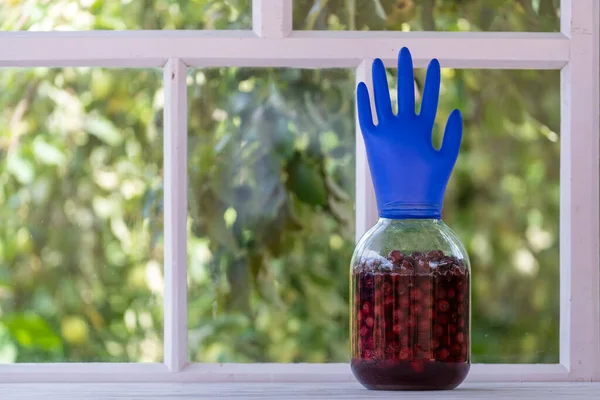 Homemade tincture of red cherry in a jar with a glove shaped shutter. Berry alcoholic drinks concept. Homemade red wine made from ripe cherries in glass jar on white wooden window background, Ukraine