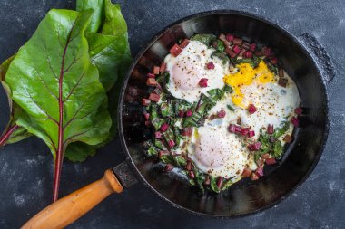 Fried eggs with green beet leaves, onion, pepper and spices in cast iron pan, close up, top view